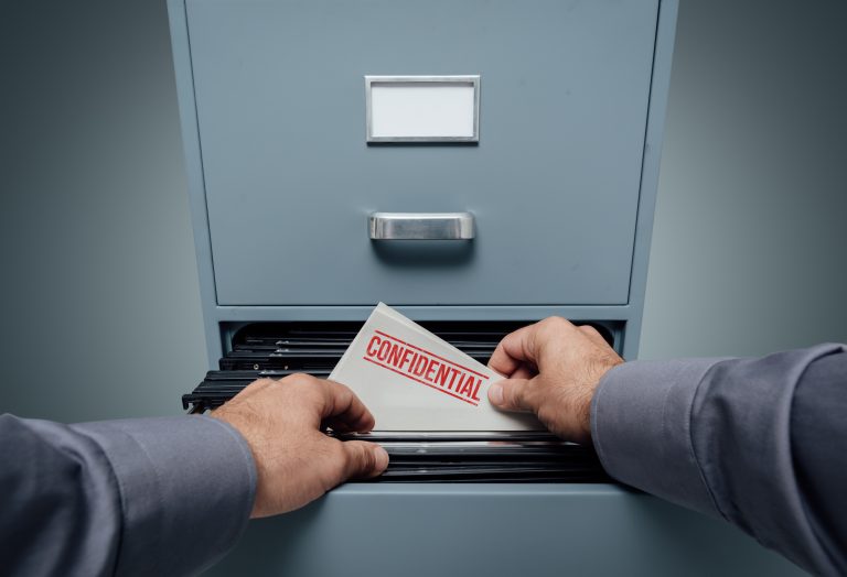 A person’s hands place an envelope labeled “CONFIDENTIAL” into a filing cabinet drawer, symbolizing the importance of privacy and discretion during the business sale process. The image emphasizes the need to protect sensitive information to ensure a smooth and secure transaction.