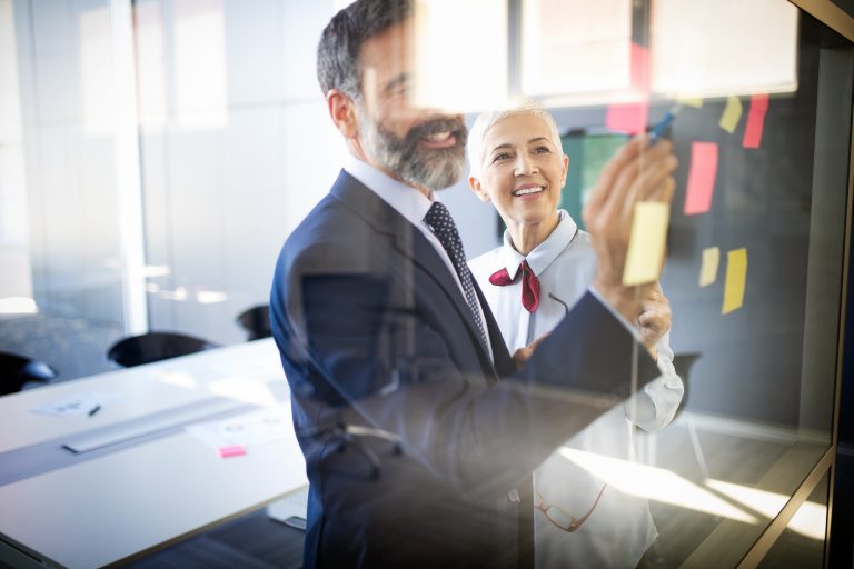 A smiling businesswoman and businessman in a modern office brainstorming together, placing sticky notes on a glass board. The image conveys collaboration, leadership, and the empowerment that comes with owning and running a business.