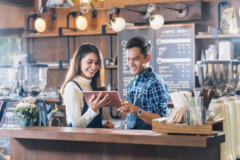 Two smiling café owners stand behind the counter reviewing information on a tablet, symbolizing teamwork, strategic planning, and business readiness. The image conveys professionalism and the importance of knowing your business's value before approaching investors.