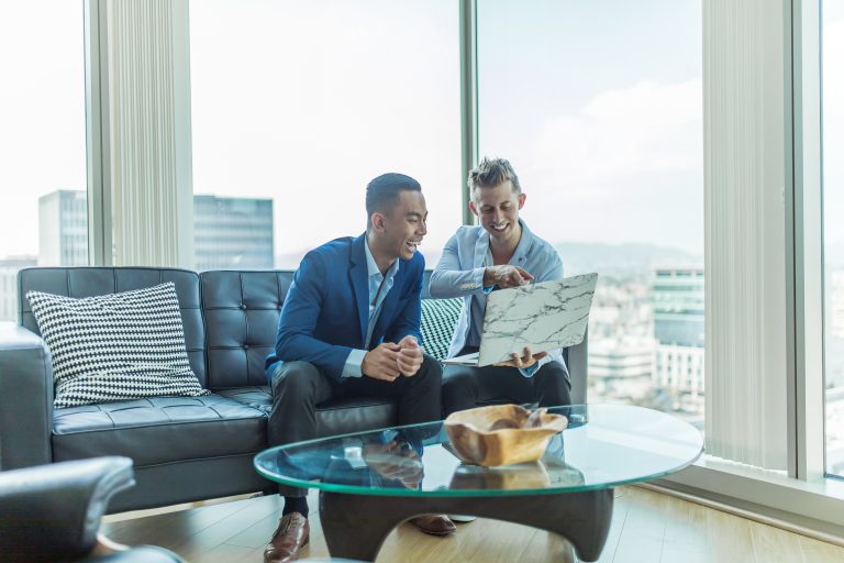 Two professionally dressed men sit on a sofa in a modern office with city views, smiling and reviewing information on a laptop. The image reflects a collaborative and confident business negotiation setting, emphasizing the importance of preparation and professionalism in deal-making.