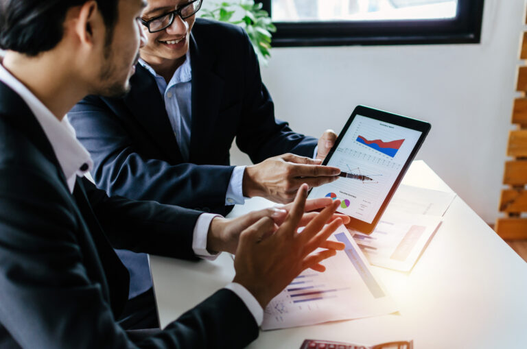 Two business professionals in suits review financial charts and graphs on a tablet during a meeting. The image reflects expert consultation, business valuation analysis, and the importance of working with a professional broker during the business sale process.