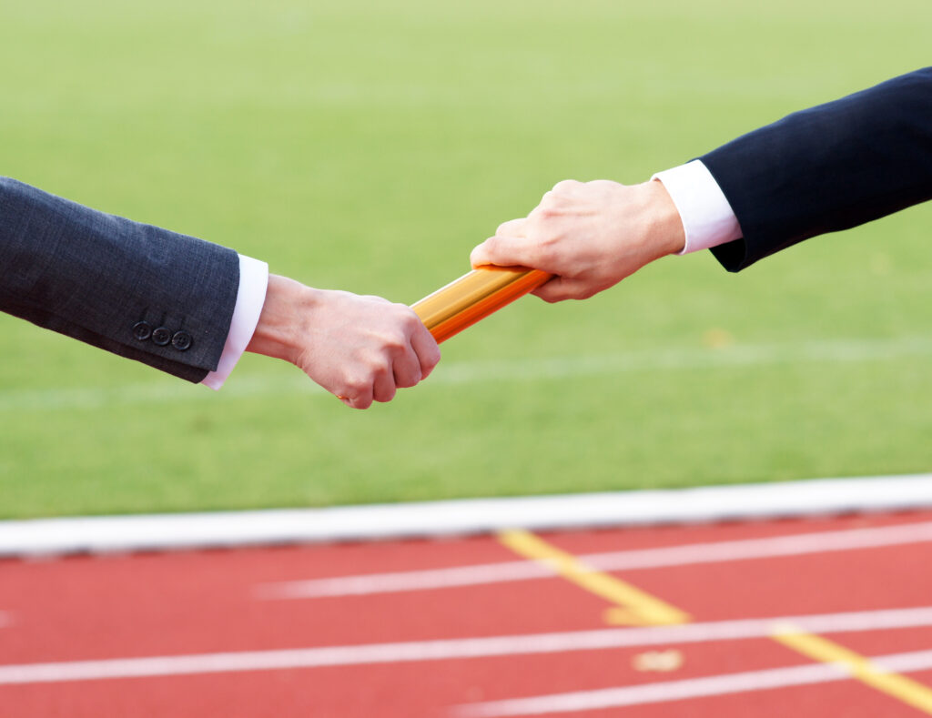 Close-up of two people in business attire passing a baton on a running track, symbolizing the smooth transition of leadership or ownership. The image represents the handoff process that occurs during a business sale and the importance of planning for continuity and long-term success.