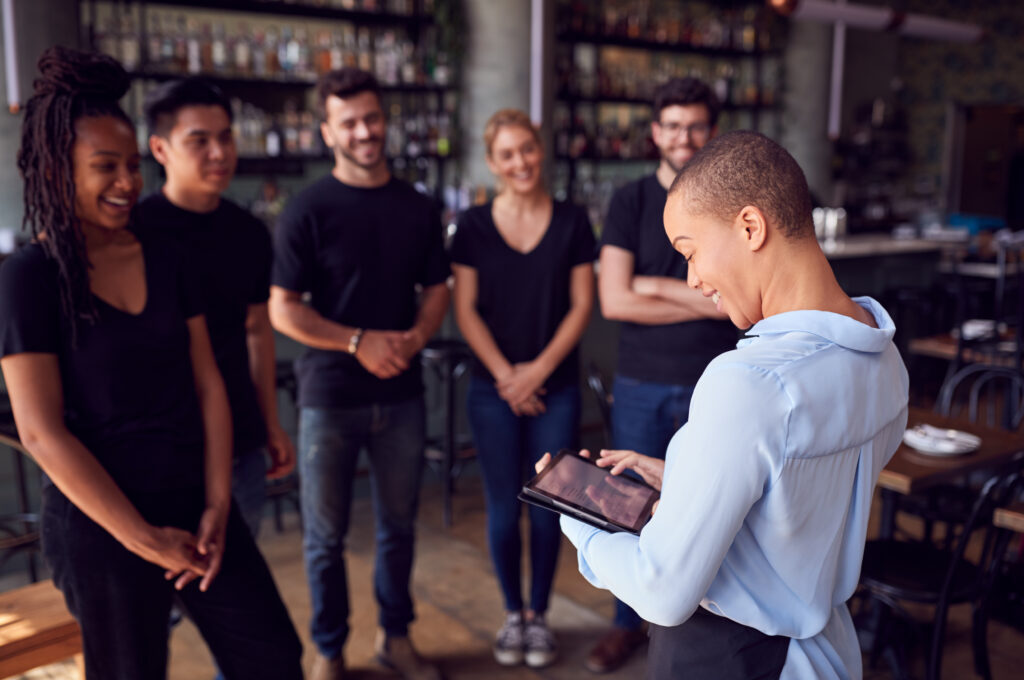A smiling business owner holding a tablet addresses a group of casually dressed employees during a team meeting inside a modern restaurant or bar. The team members appear engaged and relaxed, suggesting a positive work culture and a collaborative environment—ideal for a new owner learning the ropes post-acquisition.