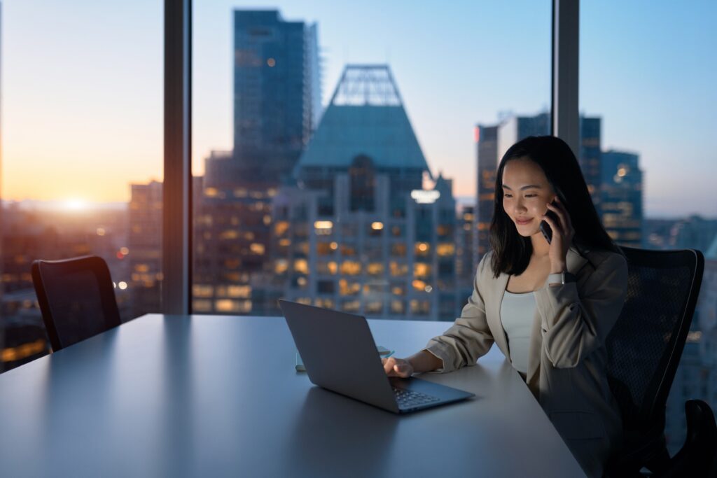 Businesswoman in a high-rise office speaking on the phone at dusk, representing a confidential business sale conversation handled discreetly and professionally.