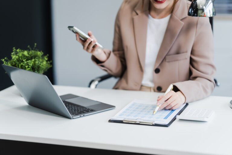 A business professional in a beige blazer reviews financial documents at a modern office desk, holding a smartphone and using a pen to analyze a printed spreadsheet, with a laptop and calculator nearby—illustrating the due diligence process before acquiring a business.