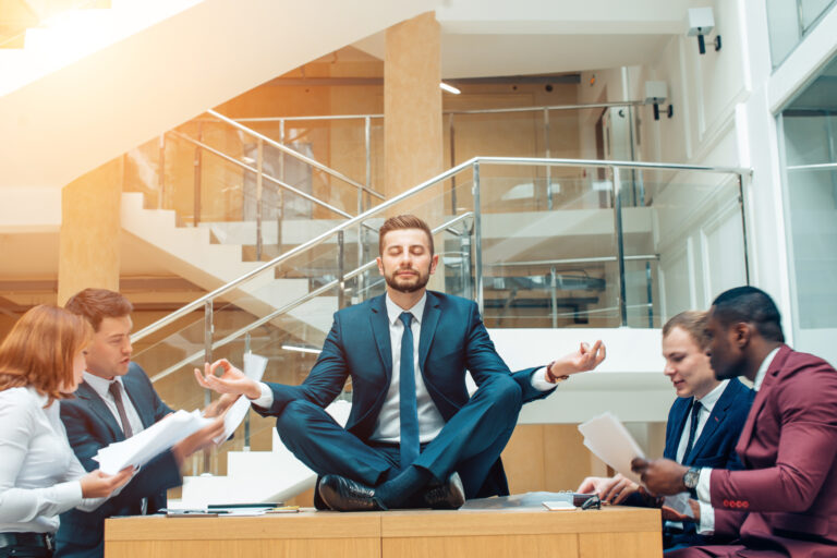 A businessman in a suit sits cross-legged atop a desk in a meditative pose, eyes closed and calm, while a team of colleagues around him frantically discuss documents—symbolizing the importance of maintaining control and calm during a business transition.