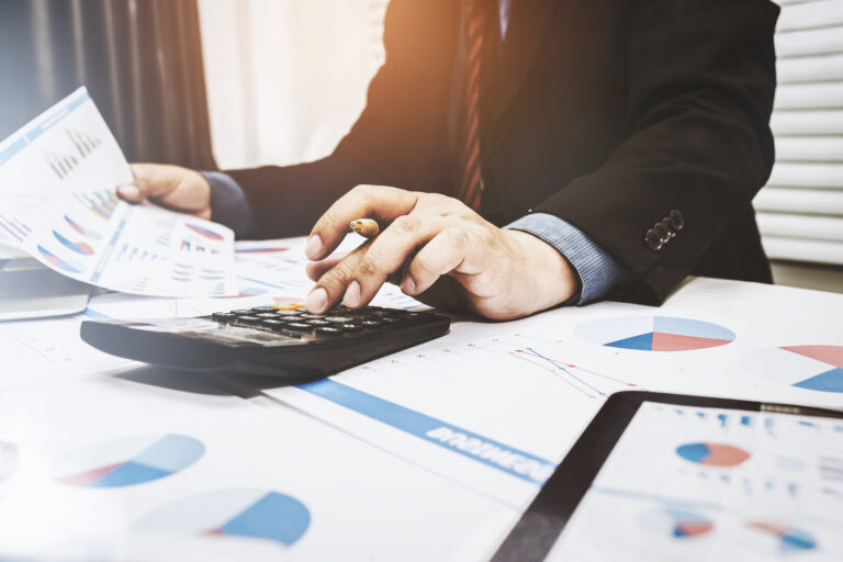 A business professional in a blazer reviews financial reports and valuation documents on a desk cluttered with a laptop, calculator, and pen—representing the analytical process of determining a business's true market value through methods like EBITDA multiples, asset valuation, and discounted cash flow.