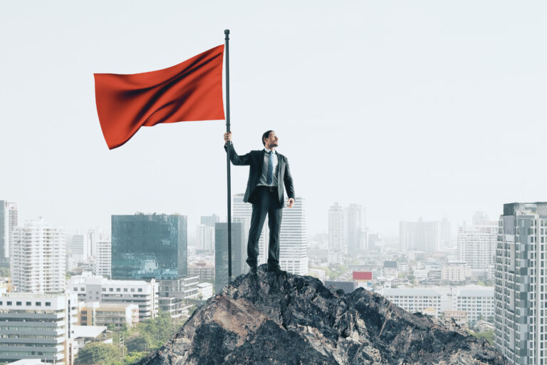 A determined businessman in a suit stands atop a rugged mountain peak, confidently holding a tall flagpole with a large red flag waving above a city skyline—symbolizing the vigilance and authority needed to spot red flags in business acquisitions.
