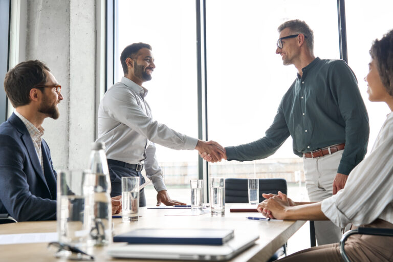 Four business professionals sit around a modern conference table during a high-stakes meeting. Two men in the center, representing buyer and seller, are captured mid-handshake—symbolizing a completed deal. Smiles and attentive expressions from the others suggest cautious optimism. Clear glasses of water and open notebooks reflect a professional setting, underscoring the importance of post-merger integration and strategic collaboration after closing.