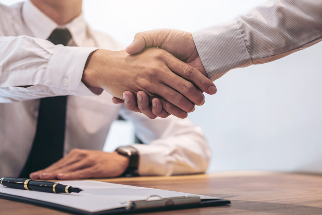Two professionals in business attire shaking hands over a finalized agreement, symbolizing a successful business acquisition. A contract, pen, and clipboard sit on the table, representing the importance of proper legal documentation in closing deals securely and strategically.