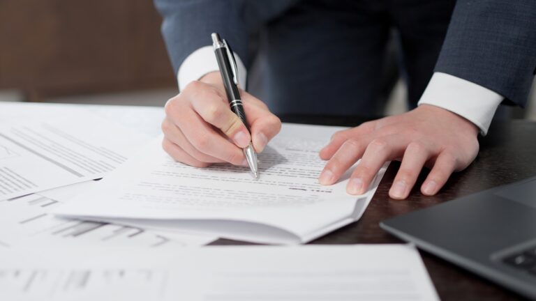 A business professional in a tailored suit signs legal documents on a desk covered with financial reports and contracts, symbolizing the complex and meticulous nature of selling a business.