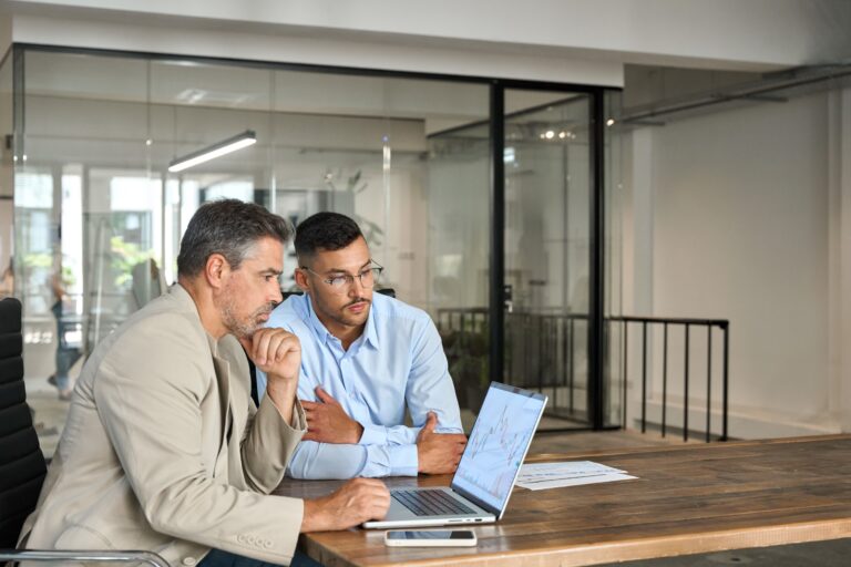 Two business professionals in a modern office setting analyze financial charts on a laptop, representing the strategic planning and expert guidance required during the complex process of buying or selling a business.