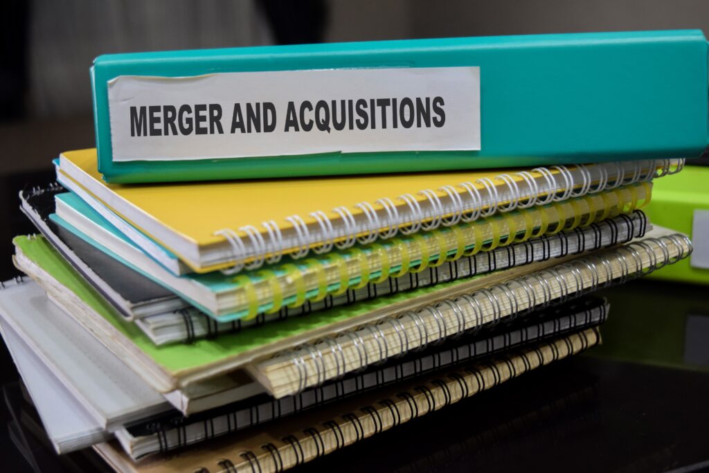Mergers and acquisitions binder stacked on business documents, representing M&A deal terms, business sales, acquisitions, earn-outs, leveraged buyouts, and transaction planning.