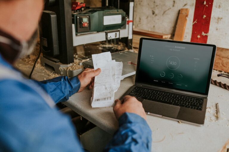 Blue-collar business owner reviewing financial records and expense receipts on a laptop inside a workshop, demonstrating financial readiness, clean bookkeeping, and operational independence to improve Freedom Score, business valuation, and exit readiness.