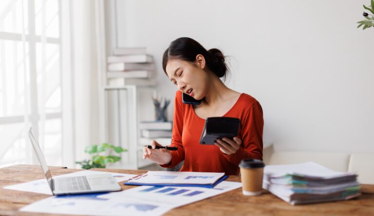 Overwhelmed small business owner multitasking with phone, calculator, and financial reports, illustrating stress from disorganized accounting and poor financial documentation during business operations and pre-sale preparation